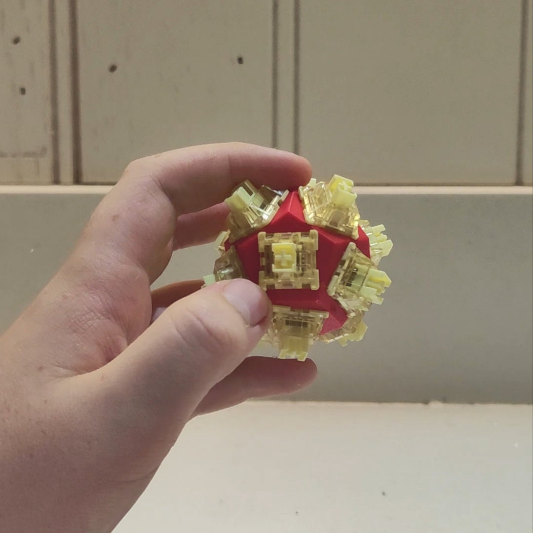 A hand holding a small red geometric puzzle ball decorated with translucent yellow switches, set against a neutral indoor background.