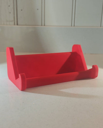 A bright red 3D-printed keyboard stand made of PLA plastic, shown on a light-colored countertop with a white paneled wall in the background.
