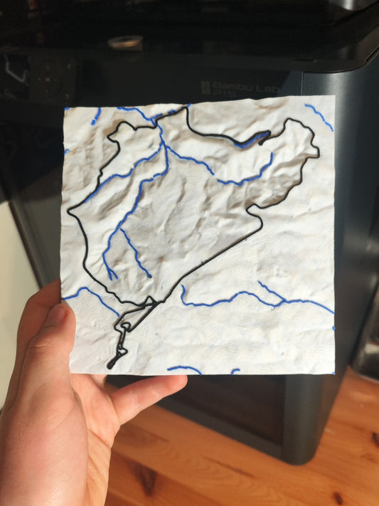 A hand holding a map design with black and blue line details, photographed in a real indoor setting with a dark appliance and wooden floor in the background.